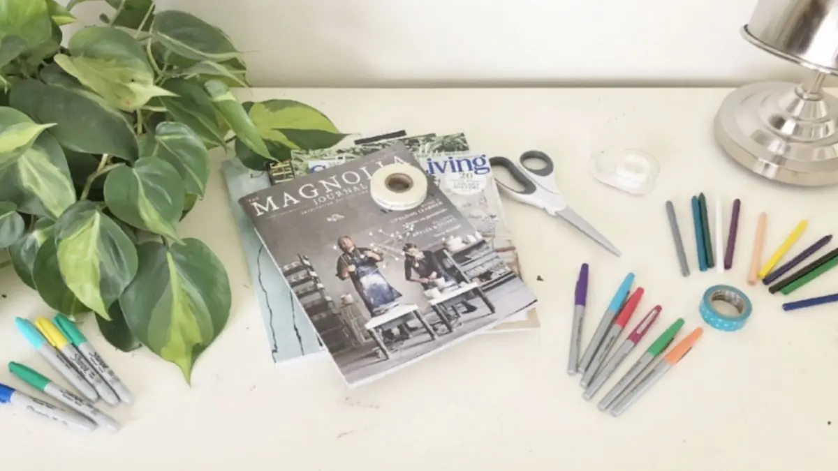 Magazines, markers, colors, and scissors next to a plant on top of a desk