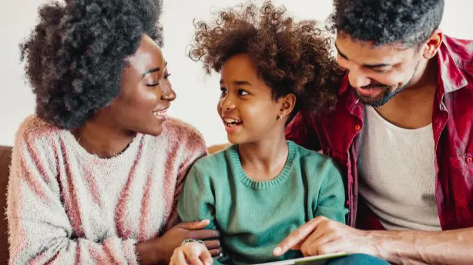 Smiling Mother and Father on each side of daughter reading with her
