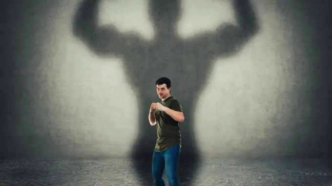 Man standing in front of wall in boxing stance while large shadow behind him is of a man flexing his muscles.
