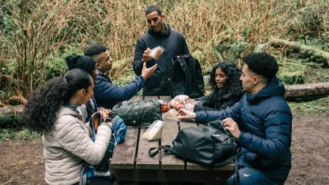 Men and women sitting around a picnic table wearing winter coats, reaching into backpacks pulling out food to share