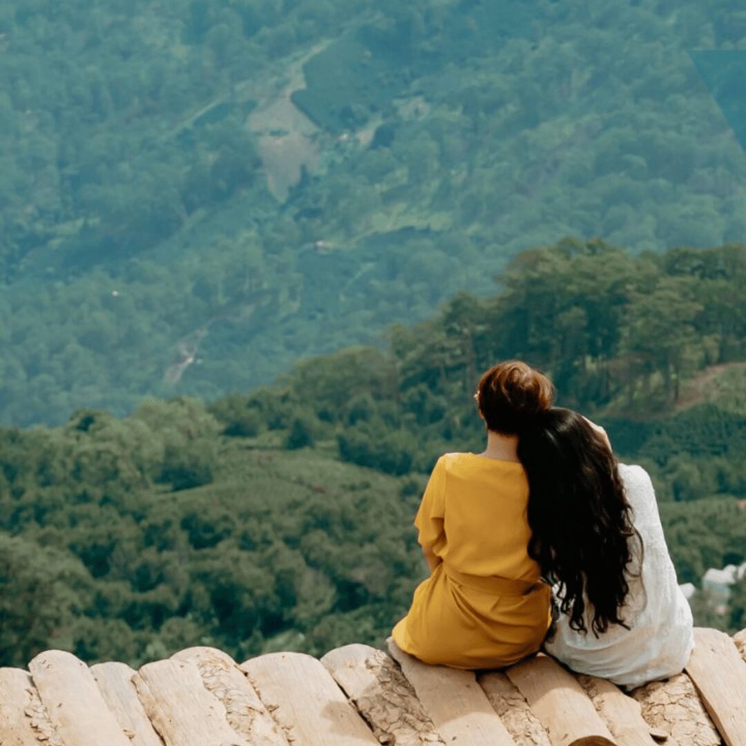 two women sitting on the edge of a cliff looking out over the forest